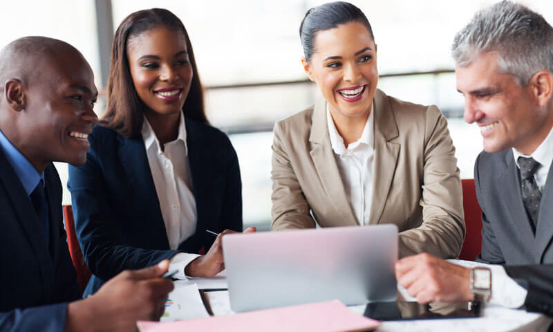 four professionals of various ethnicities sitting around a table discussing what they see on a laptop